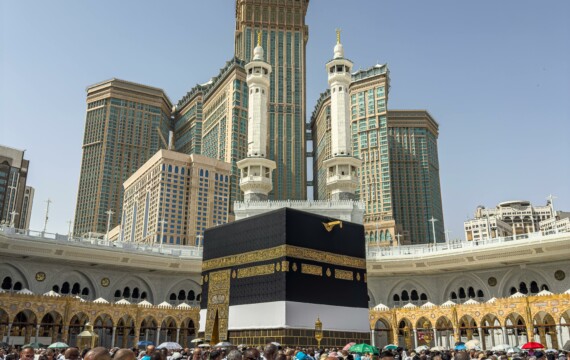 A large group of pilgrims gather at the Kaaba in Mecca, Saudi Arabia, during the Hajj pilgrimage under a clear blue sky.
