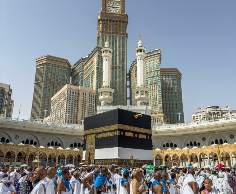 A large group of pilgrims gather at the Kaaba in Mecca, Saudi Arabia, during the Hajj pilgrimage under a clear blue sky.