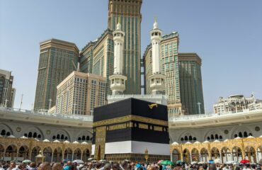 A large group of pilgrims gather at the Kaaba in Mecca, Saudi Arabia, during the Hajj pilgrimage under a clear blue sky.