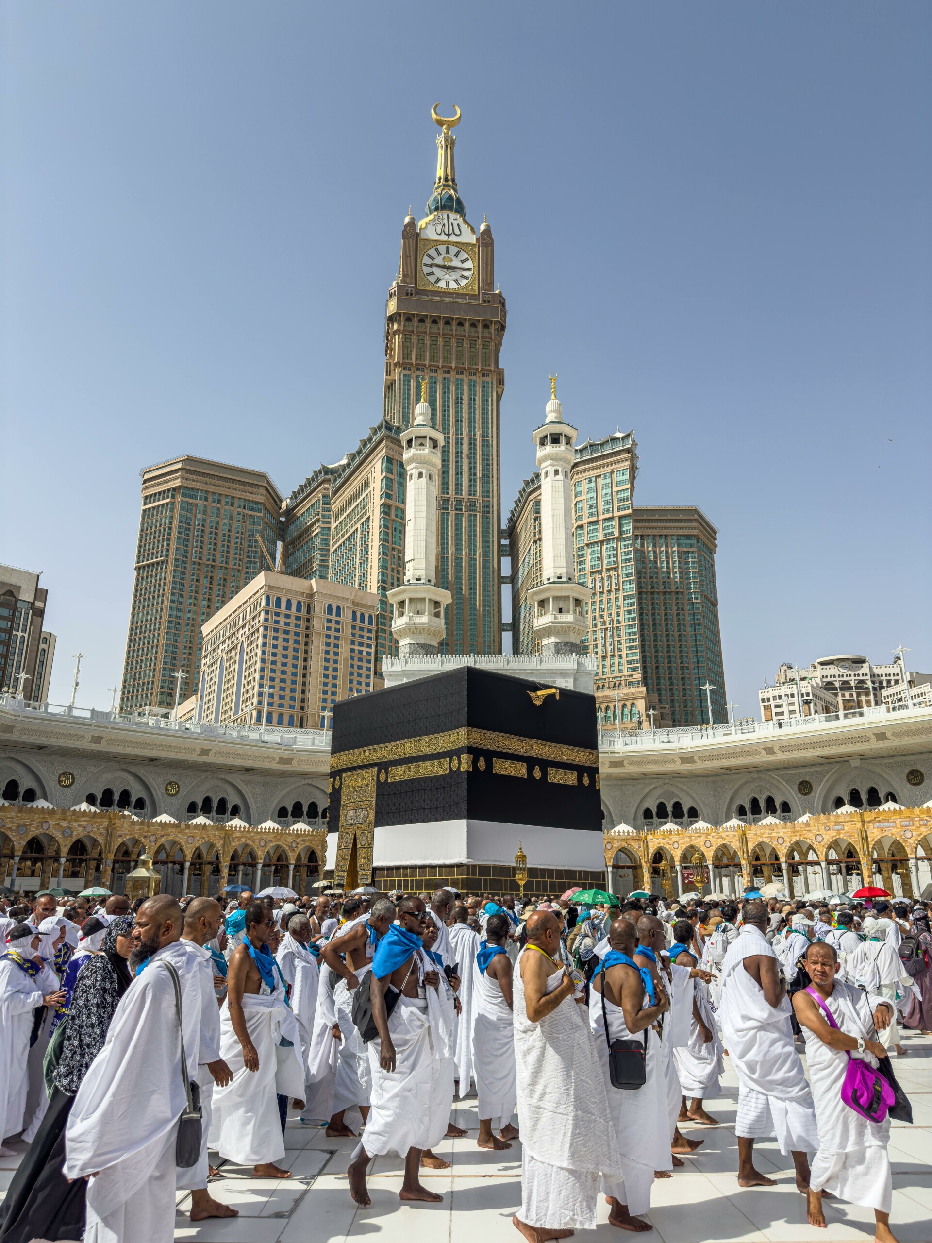 A large group of pilgrims gather at the Kaaba in Mecca, Saudi Arabia, during the Hajj pilgrimage under a clear blue sky.